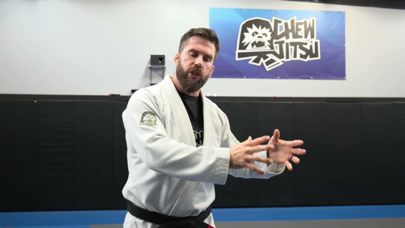 A black belt in a white gi stands on the mats explaining a lesson.
