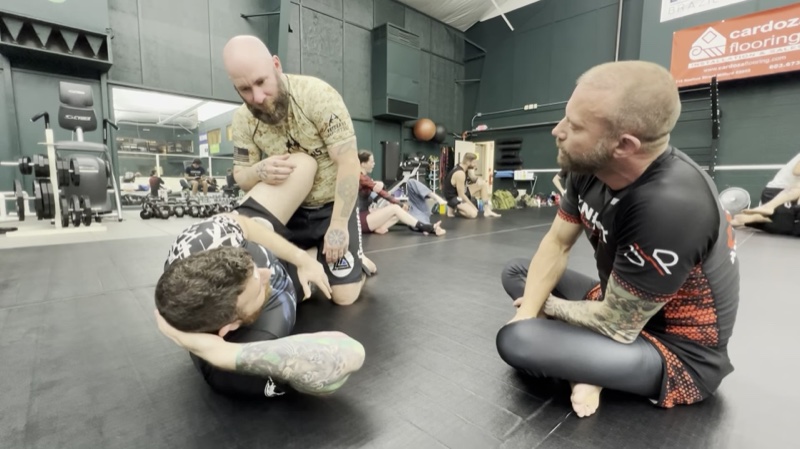 An instructor sits beside two students as they drill, with other pairs training in the background.