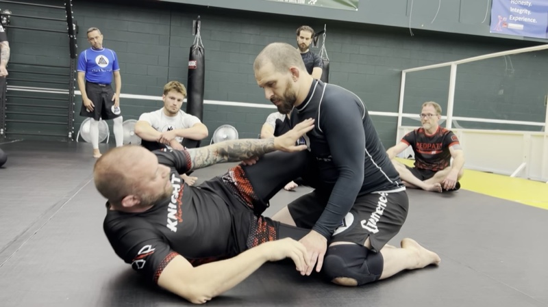 An instructor on his back demonstrates a hold on a kneeling partner while students sit and watch.