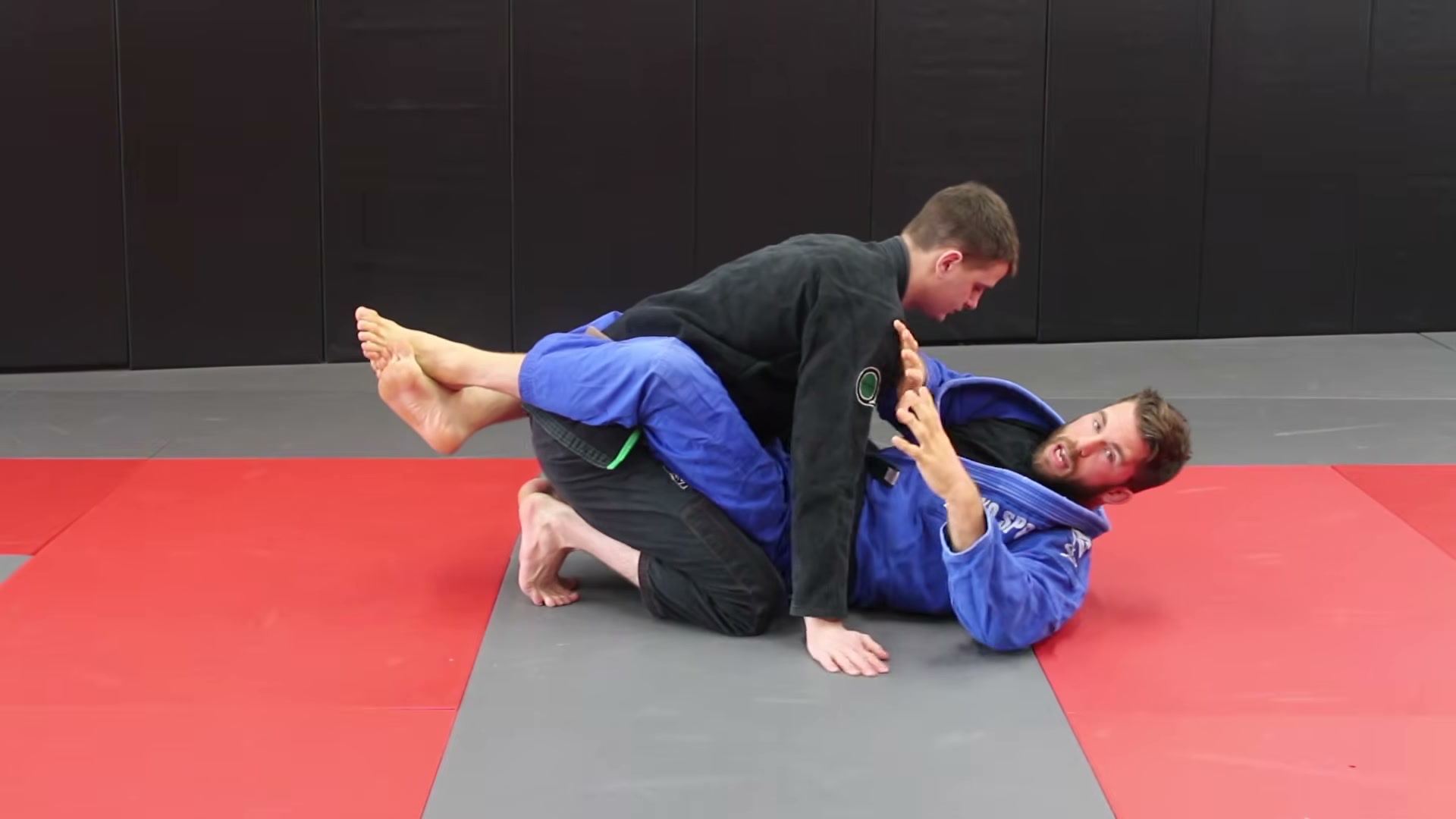The bottom player in a blue gi locks closed guard around a kneeling partner in a black gi and talks through a detail.