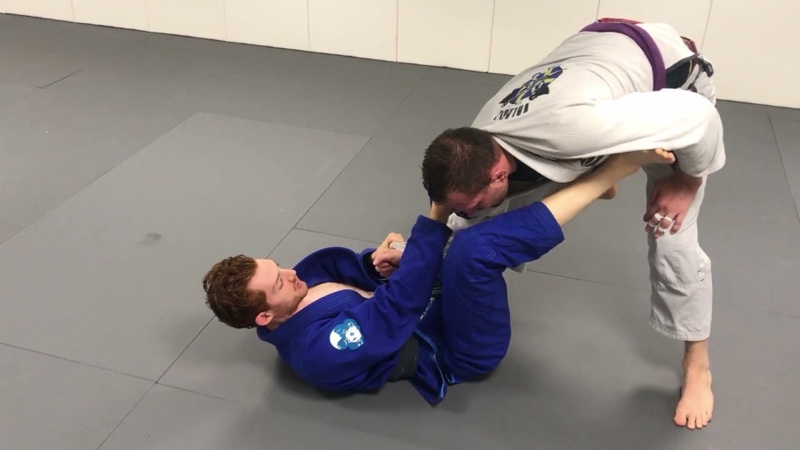 The bottom player in a blue gi holds a standing opponent's collar and sleeve, with a foot on the biceps, in collar-sleeve guard.