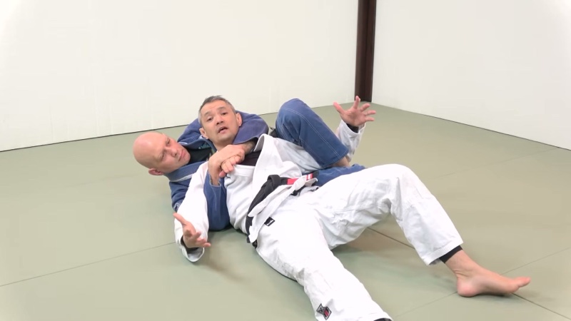 The attacker in a blue gi is perpendicular behind the opponent in a white gi, with one arm trapped between the legs, in the crucifix.