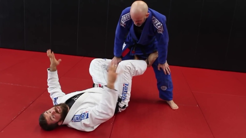The bottom player in a white gi hooks the standing opponent's lead leg from the outside in De La Riva guard.