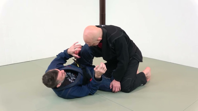 The bottom player in a blue gi traps one of the kneeling opponent's legs between his own in half guard.