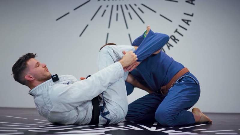 The bottom player in a white gi wraps a shin around the kneeling opponent's arm and holds the sleeve in lasso guard.