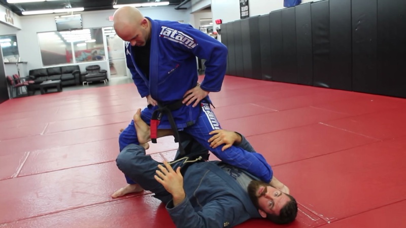 The bottom player lies underneath a standing opponent, legs crossed around one thigh in X-guard.