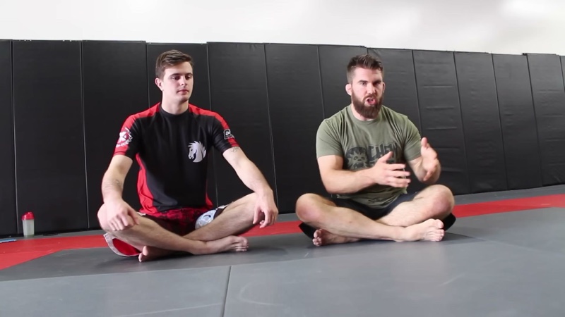An instructor sits on the mats next to a student and explains a point with his hands.